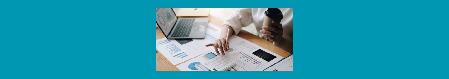 Services Services
Blue background, light colored wood desk, open lap top with charts, papers with different chars, white calculator, a woman's torso in white shirt sleeves rolled up, cup of coffee ( brown to go cup with white lid, in left hand, pencil in the right.