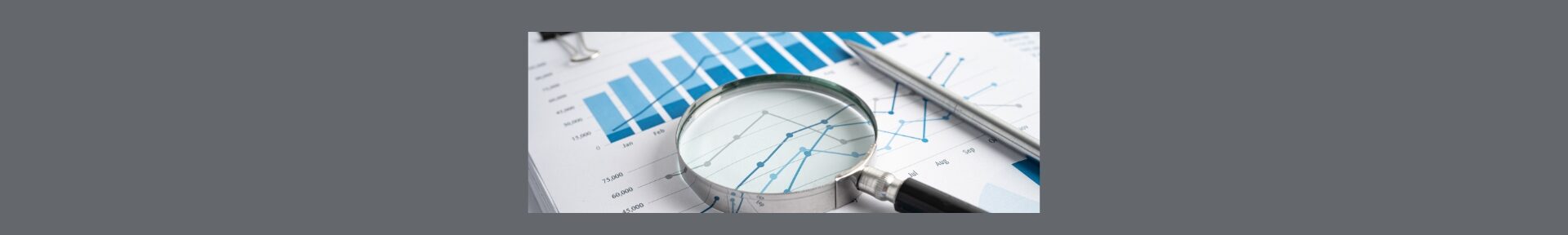 Understanding Financial Report Terminology A Glossary for Beginners. Dark gray background, with a white block in the middle of a white sheet of paper with graphs and a silver pen, a silver magnify glass with black handle.