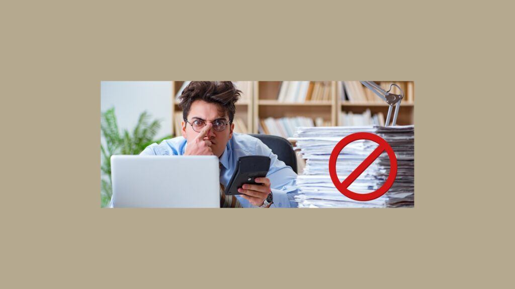 How to Avoid Falling Behind on Your Bookkeeping Again
Light brown bookshelves in the background. Man in dress shirt, right pointer finger on nose piece on glasses, left hand holding a calculator, open laptop in front and a stack of papers on the right with a large red circle with a red line through the center.